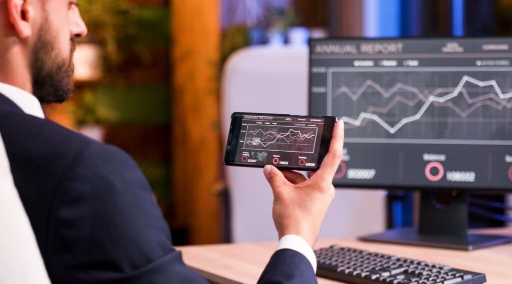 Businessman holding a phone displaying stock charts, with a digital screen chart in the background